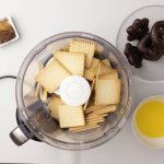 Butterkekse im Küchenmixer, daneben Schoko-Brezeln, brauner Zucker und geschmolzene Butter