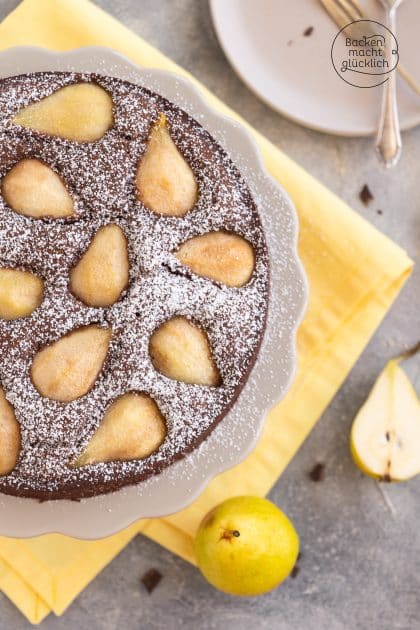 Schoko-Birnenkuchen mit Puderzucker bestäubt, ganze Birnen daneben auf gelbem Tuch