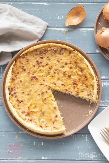 Angeschnittener Zwiebelkuchen mit Speckwürfeln auf rundem Backblech, Zwiebeln im Hintergrund