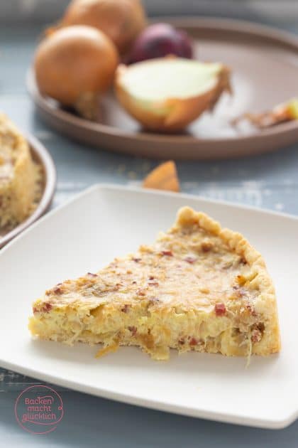 Zwiebelkuchenstück auf weißem Teller, dahinter Zwiebeln auf braunem Servierteller