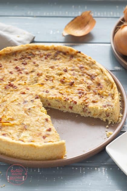 Angeschnittener Zwiebelkuchen mit Mürbeteig auf Teller, Zwiebeln im Hintergrund