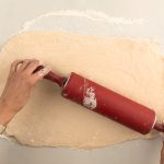 Hefeteig wird mit rotem Nudelholz auf bemehlter Arbeitsfläche ausgerollt