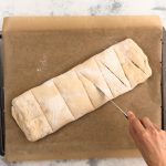 Hand schneidet Einschnitte in gerollten Handbrot-Teigling auf Backpapier