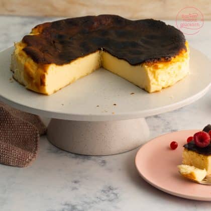 Angeschnittener San Sebastian Cheesecake mit karamellisierter Kruste auf Kuchenständer, Stück auf rosa Teller