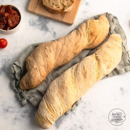Zwei goldbraune Wurzelbrote auf grauem Tuch, daneben getrocknete Tomaten und Kirschtomate