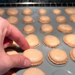 Hand prüft frisch gebackene Macaronschalen auf Silikonbackmatte im Ofen